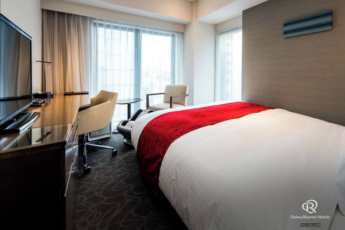 Una habitación de hotel con cama, escritorio, silla, televisor y ventana con cortinas.