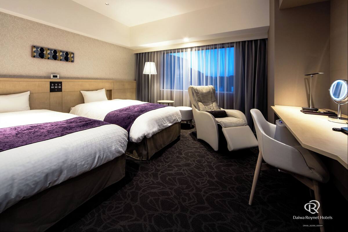 Habitación de hotel con dos camas, un escritorio, una silla, una lámpara y una ventana con cortinas.