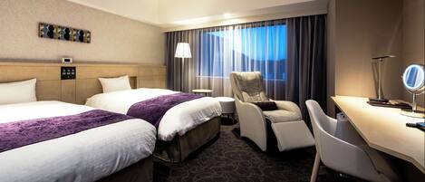 A hotel room with two beds, a desk, a chair, a lamp, and a window with curtains.