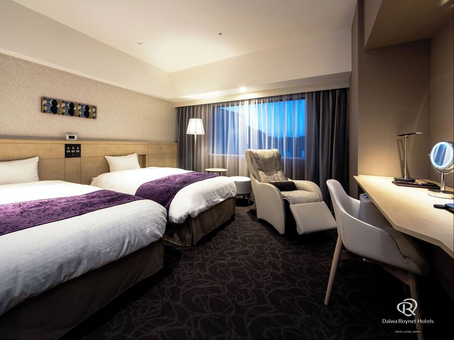 A hotel room with two beds, a desk, a chair, a lamp, and a window with curtains.