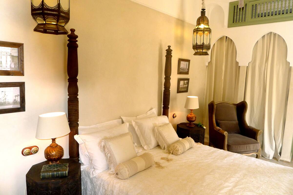 A neatly made bed with white linens and multiple pillows, a wooden nightstand with a lamp, a chair, and framed pictures on the wall.
