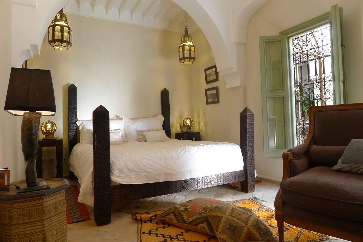 A bedroom with a four-poster bed, a nightstand with a lamp, a decorative floor rug, and a window with a metal screen.