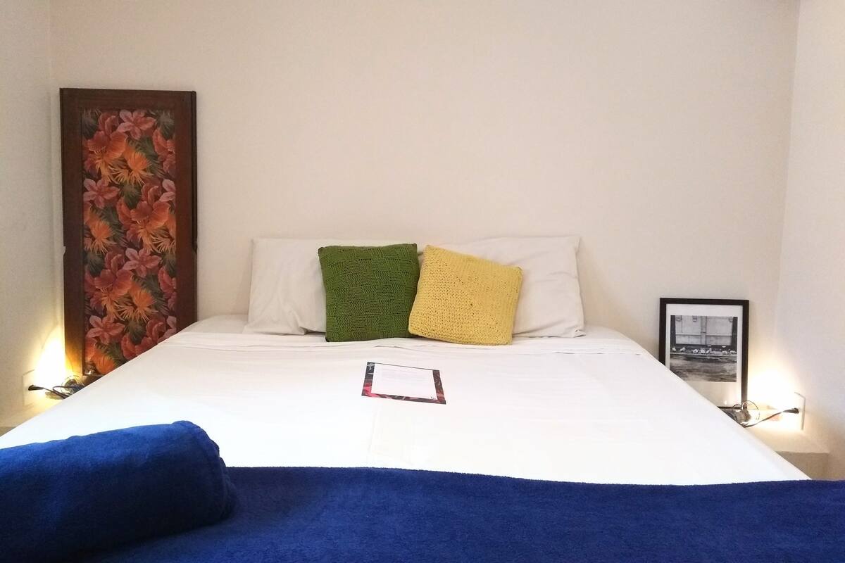A neatly made bed with white sheets, a blue blanket, and three pillows in green, yellow, and white. There is a framed picture on the wall to the right and a large floral artwork on the left.