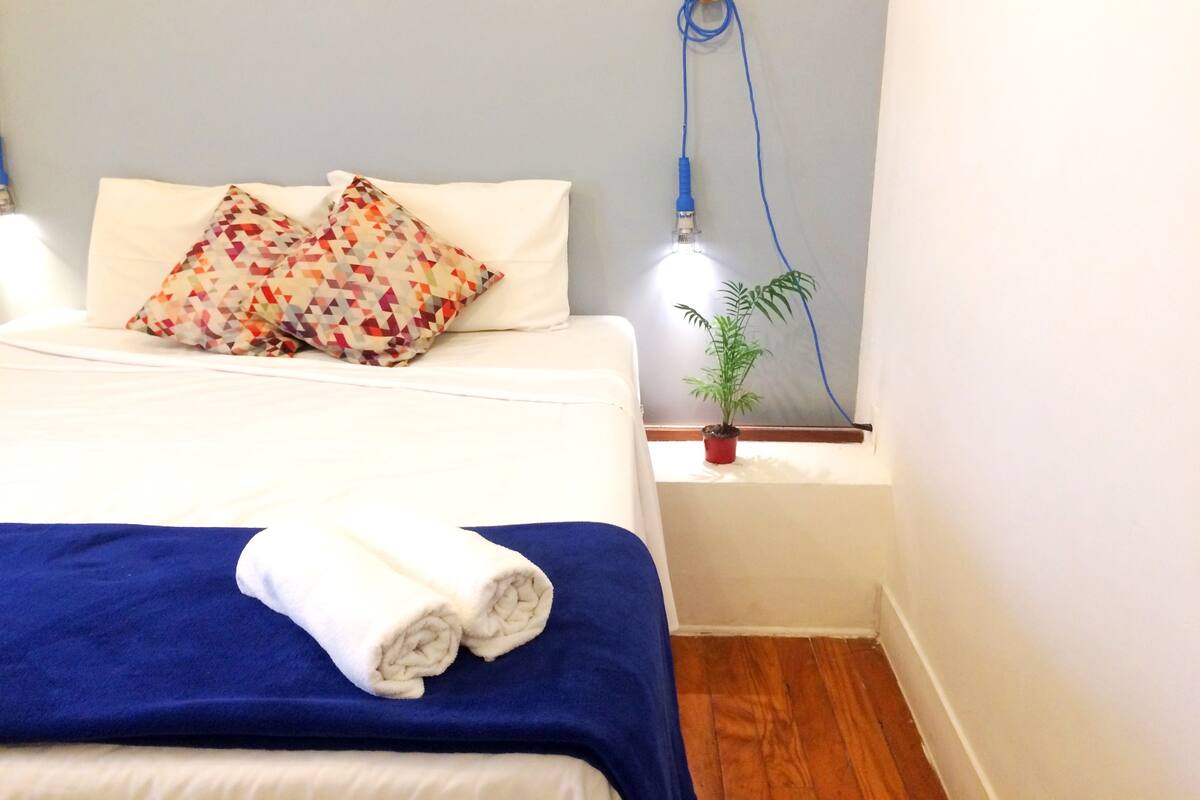 A neatly made bed with a blue blanket, two rolled towels, and a colorful pillow. There is a small potted plant on a bedside table.