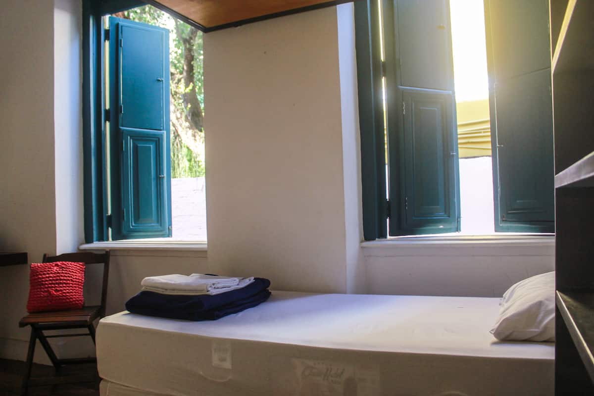 A small room with a bed, a chair, and a window with open shutters.