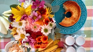 Una mesa con diversos elementos, incluyendo un plato con un trozo de pastel, un tazón de mandarinas, un racimo de plátanos, varias tazas y un arreglo floral.