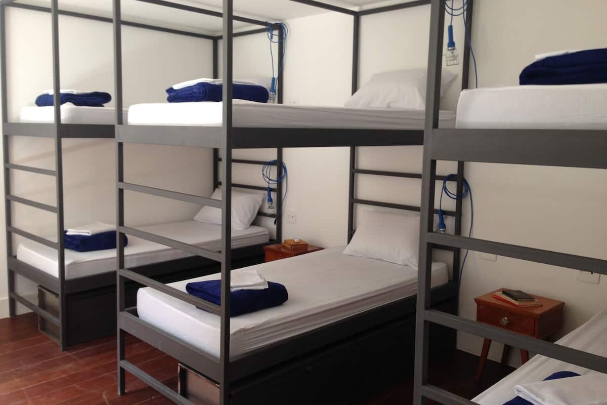 A bunk bed room with three beds, each with a pillow and a blanket. There is a wooden nightstand with a book and a lamp.