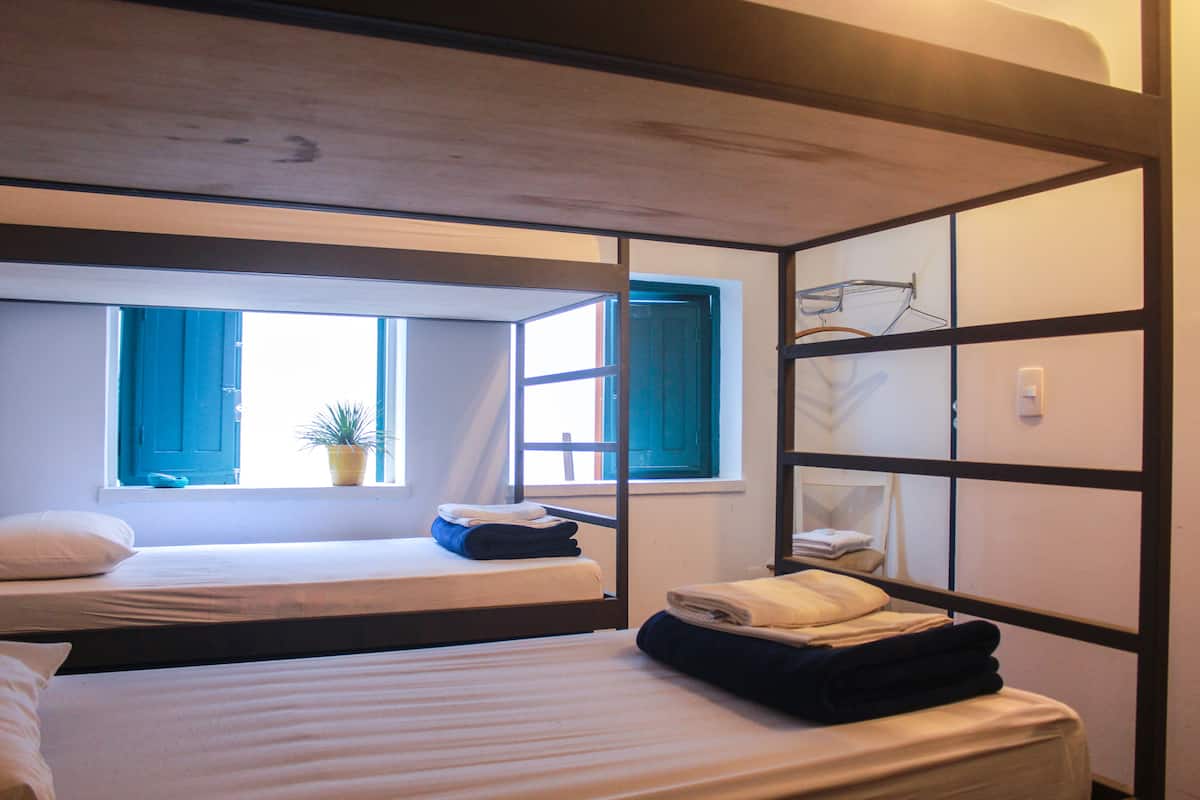 A dormitory-style room with bunk beds, a window with blue shutters, a potted plant, and a shelf with folded towels.