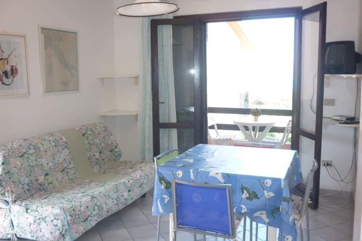 Una habitaci ón con un sofá floral, una mesa con mantel azul, una lámpara de techo y vistas a través de un ventanal.
