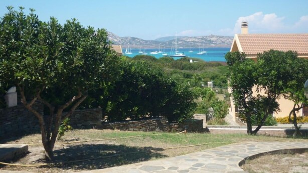 A stone pathway leading to a building with a tiled roof, surrounded by greenery and a view of boats on the water.