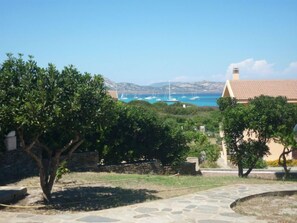 Um caminho de pedra leva a um edifício com telhado em azulejos, rodeado por vegetação e com vista para barcos na água.