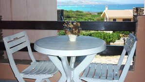 A balcony with a round table and two white chairs, overlooking a coastal view with buildings and a clear sky.