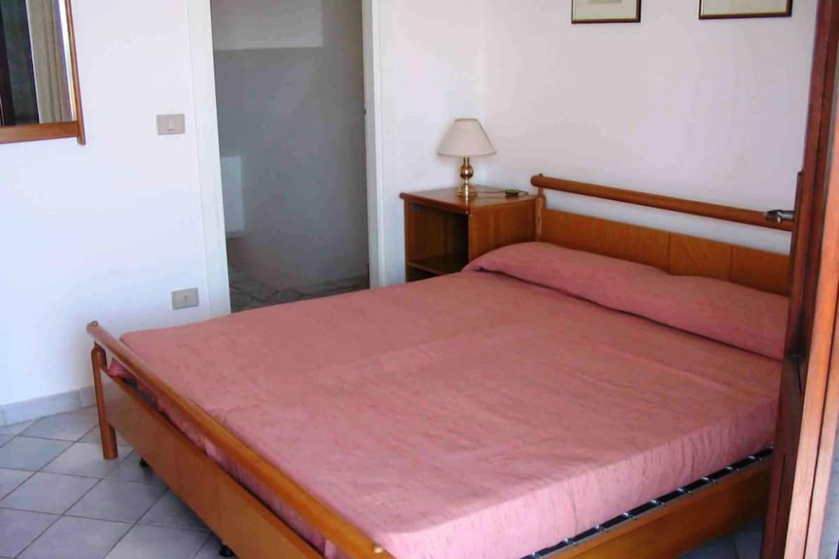 Un dormitorio con una cama de madera, una mesita de noche con una lámpara y un pequeño marco de fotos en la pared.