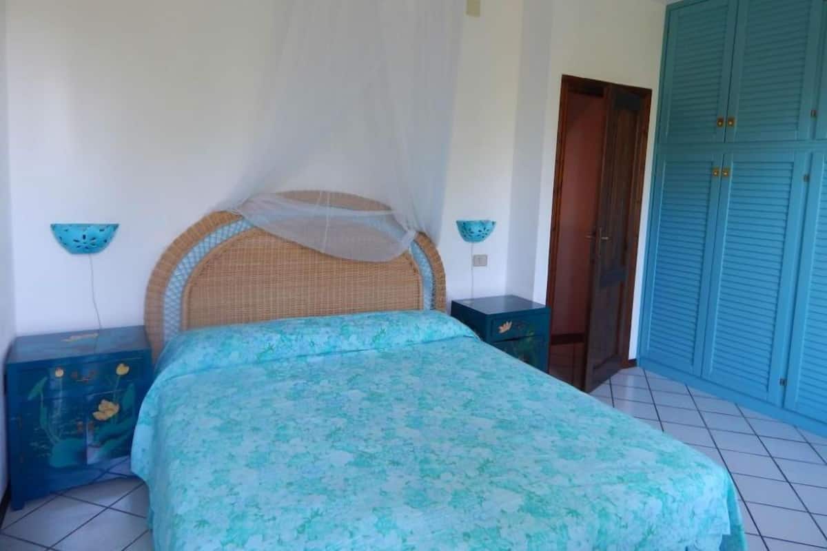 Un dormitorio con una cama, mosquitera, un armario azul y una mesita de noche con un objeto decorativo.