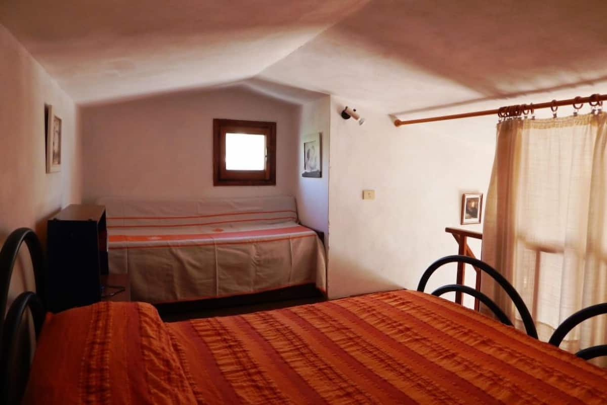 A small, single-room accommodation with a bed, a chair, a desk, and a window with curtains.