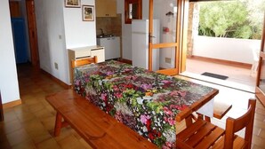 A room with a wooden table and bench, a bed with a floral bedspread, a kitchenette, and a view through a glass door. - AbitaSardegna Pittulongu (Olbia)