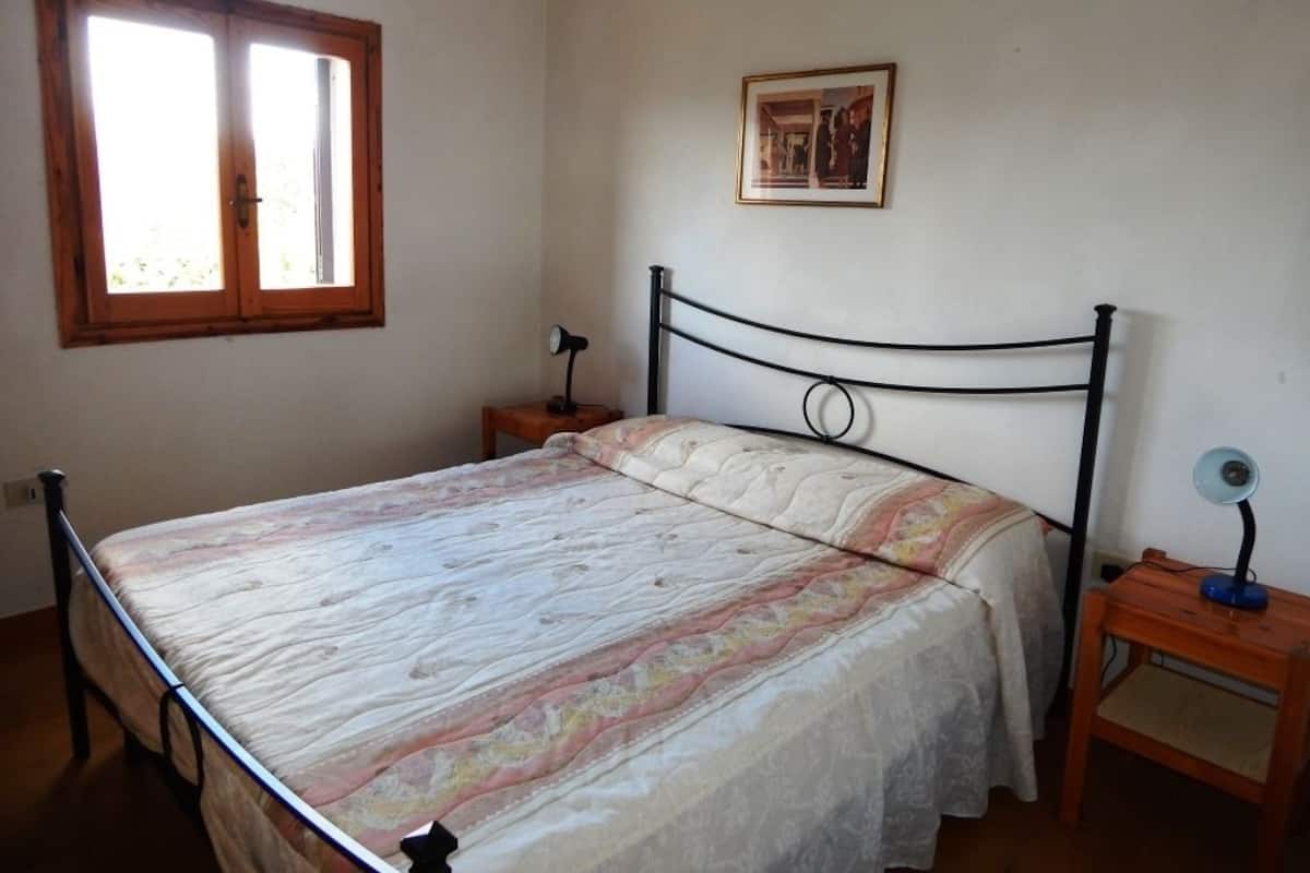 A bedroom with a bed, a window, a bedside table, a lamp, and a framed picture on the wall.