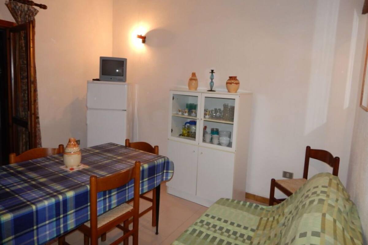 A small, cozy room with a dining table, chairs, a bed, a cabinet with glass doors, and a television on a stand.