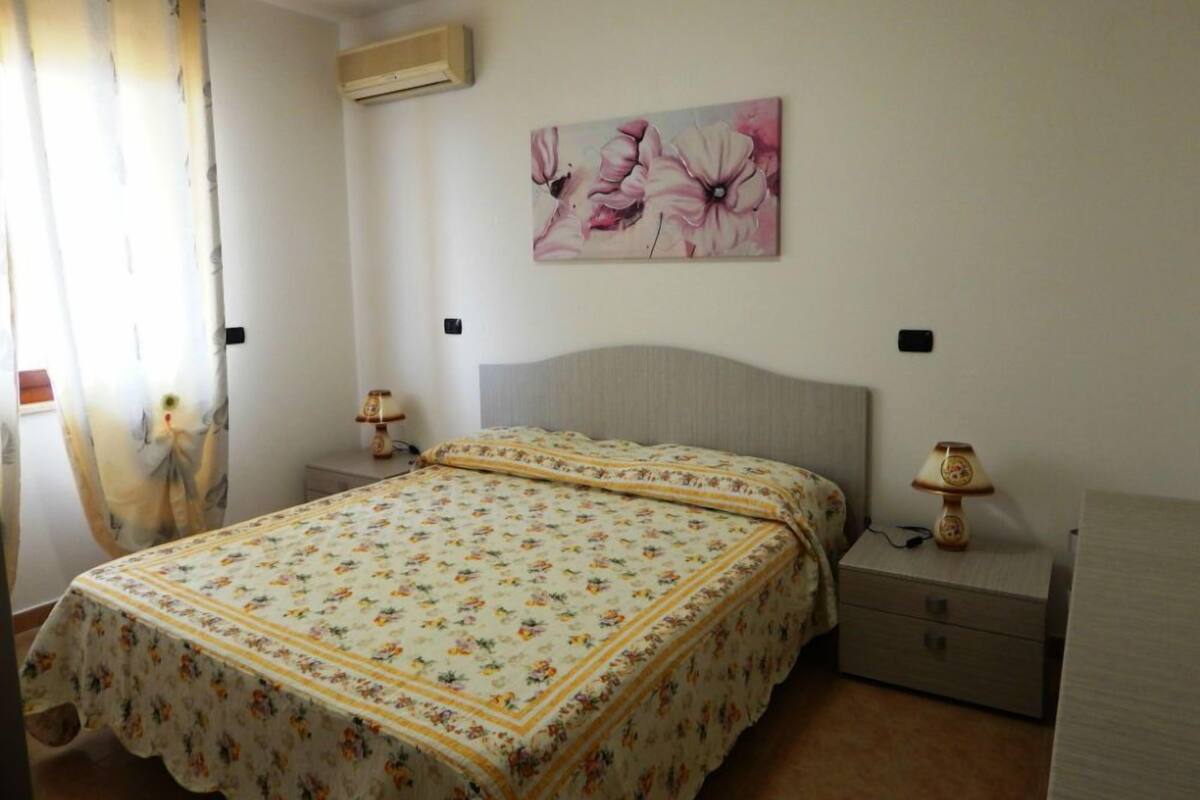 A bedroom with a floral bedspread, a headboard, two bedside tables with lamps, and a wall-mounted air conditioner.