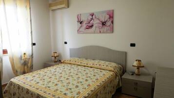 A bedroom with a floral bedspread, a headboard, two bedside tables with lamps, and a wall-mounted air conditioner.