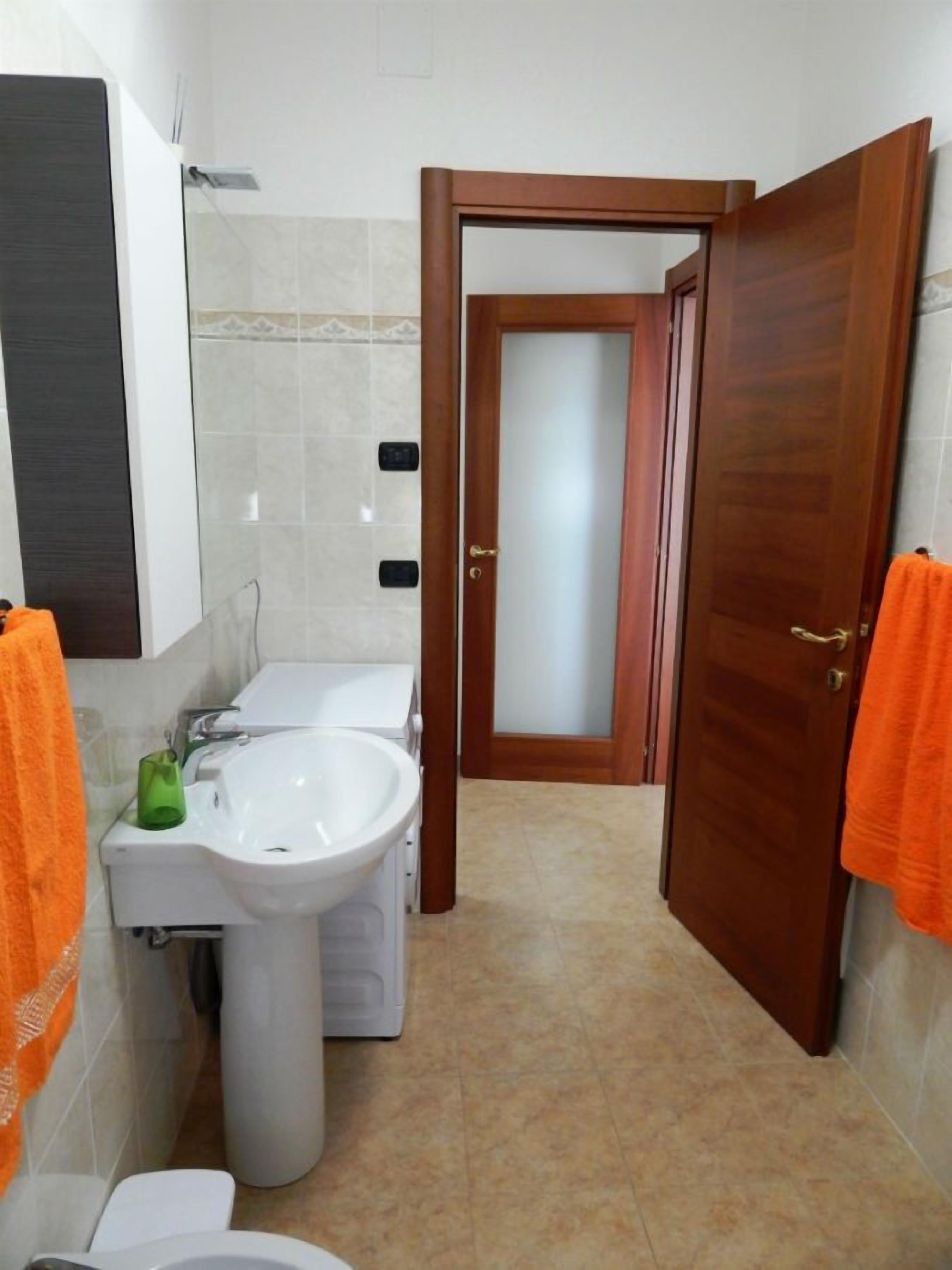 Un bagno con servizi igienici, lavandino e specchio. Si vede una porta che conduce in un'altra stanza.