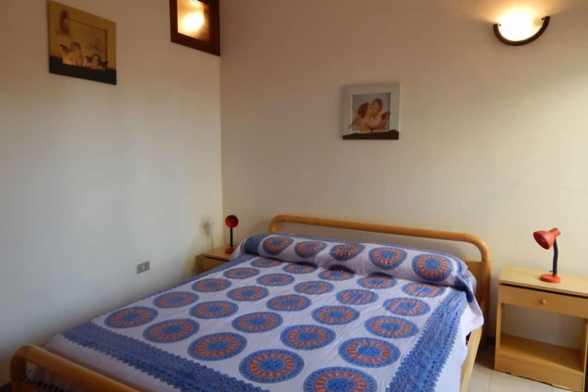 A bedroom with a bed, a nightstand, a wall-mounted lamp, and two framed pictures.