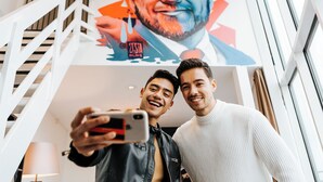 Twee personen maken een selfie in een moderne ruimte met een trap en een groot muurschildering op de achtergrond.