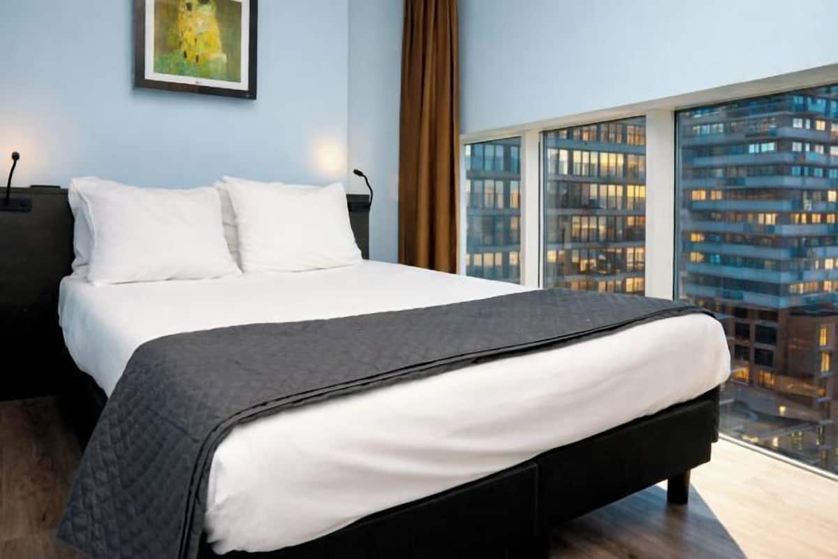 Une chambre moderne avec un grand lit, une vue sur la ville et une œuvre d’art encadrée au mur.