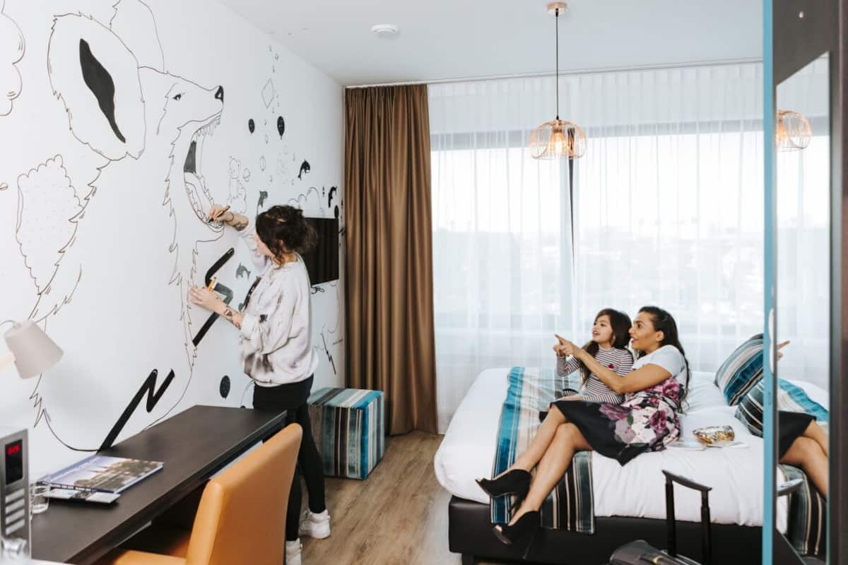 Une chambre d’hôtel avec une grande fresque murale, un lit avec une literie à rayures, un bureau avec un ordinateur portable et une fenêtre avec des rideaux.