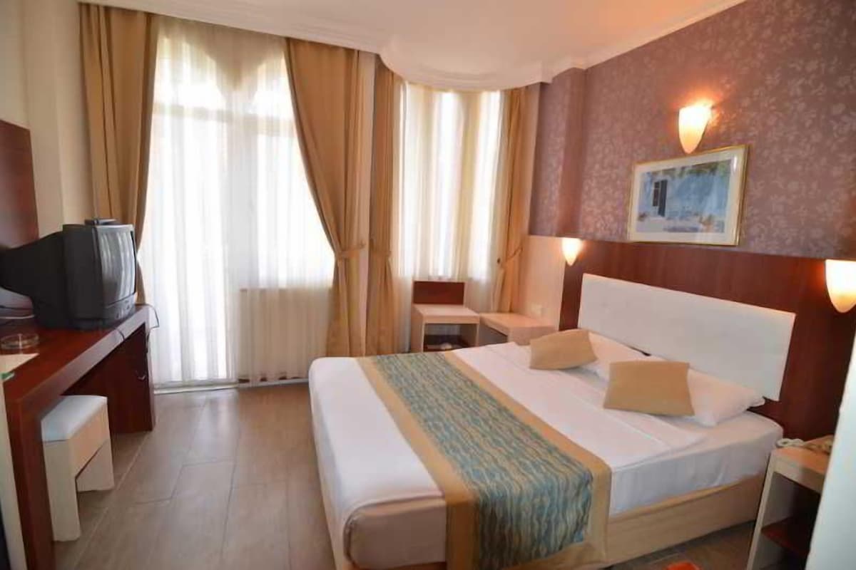A hotel room with a bed, a desk with a television, a chair, a bedside table, and a lamp.