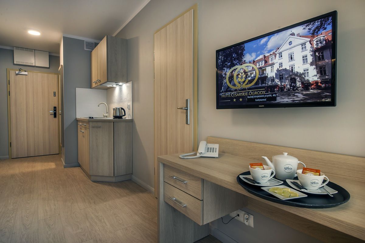 Une chambre d’hôtel équipée d’une kitchenette, d’un bureau avec un téléphone et une théière, et d’un téléviseur fixé au mur qui affiche une image de l’hôtel.