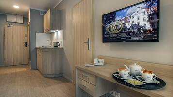 Une chambre d’hôtel équipée d’une kitchenette, d’un bureau avec un téléphone et une théière, et d’un téléviseur fixé au mur qui affiche une image de l’hôtel.