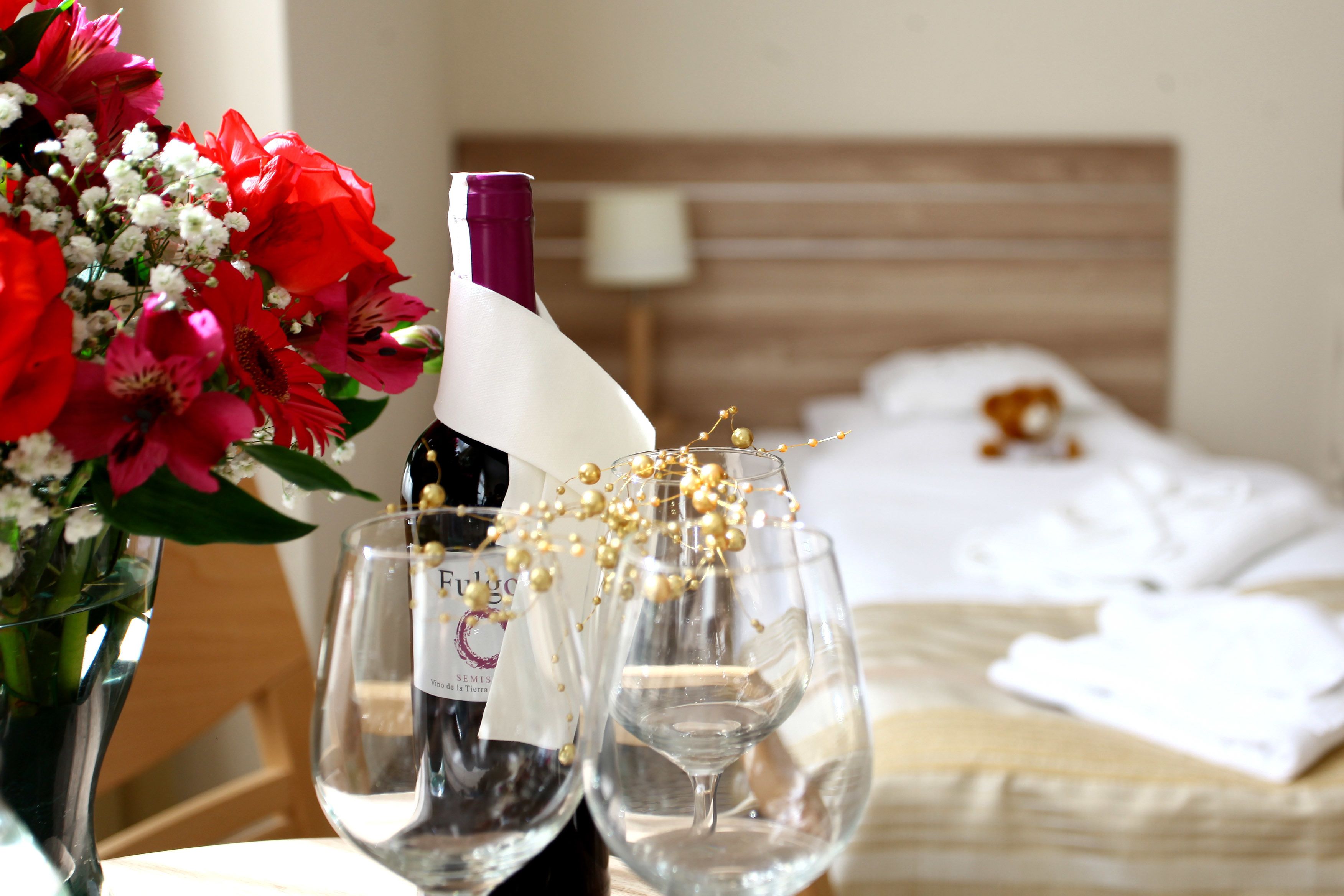 A hotel room with a bed, a bedside table with a vase of flowers, two wine glasses, and a bottle of wine.