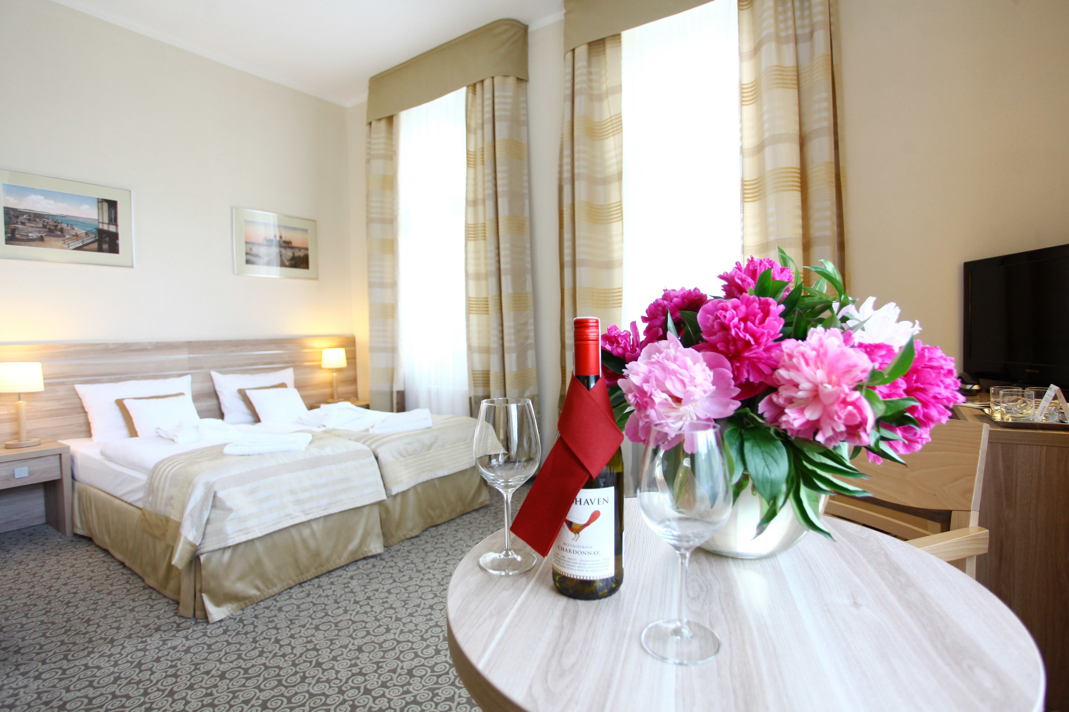 Una habitación de hotel con una cama, una mesa con una botella de vino, una copa y un jarrón con flores.