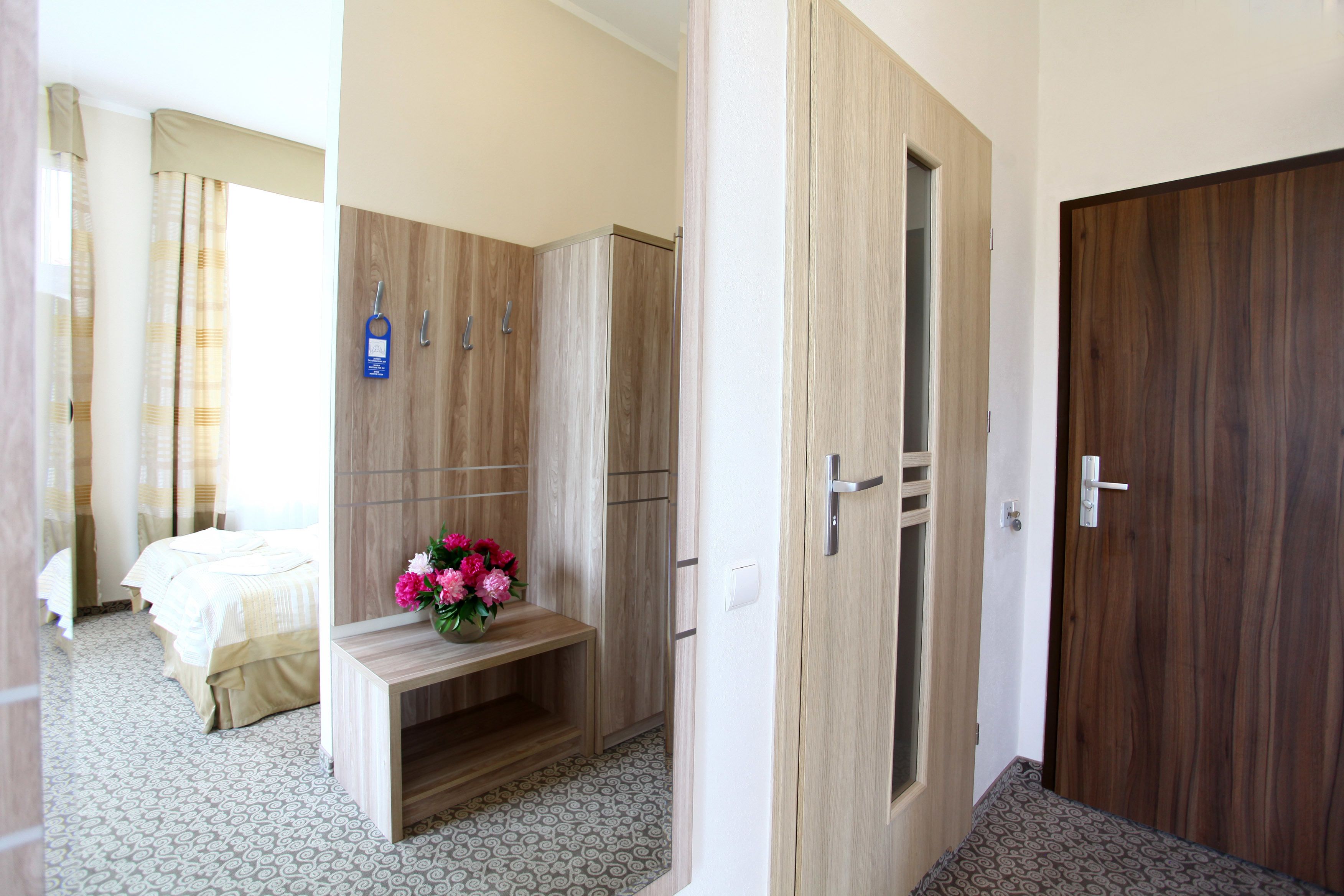 A hotel room with a bed, a wooden wardrobe, a bench with flowers, and a door.