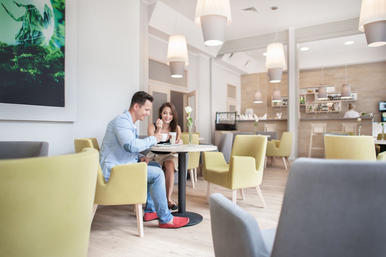 A modern hotel lobby with yellow chairs, a round table, and a bar area in the background.