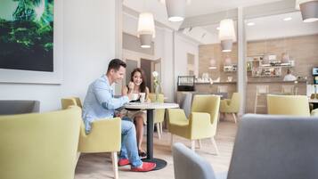 A modern hotel lobby with yellow chairs, a round table, and a bar area in the background.