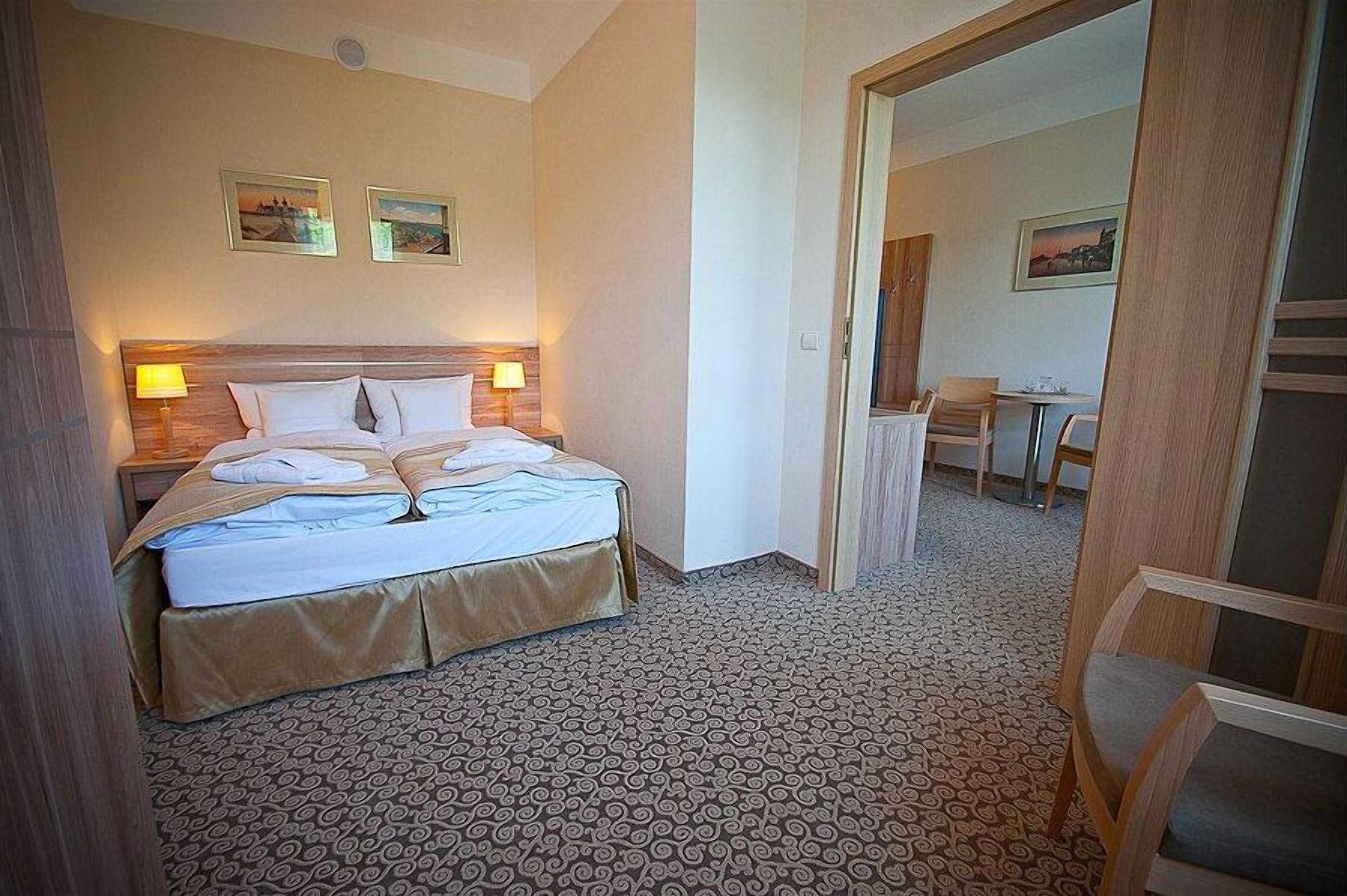 Una habitación de hotel con cama, dos lámparas de noche, una pequeña mesa con sillas y piso alfombrado.