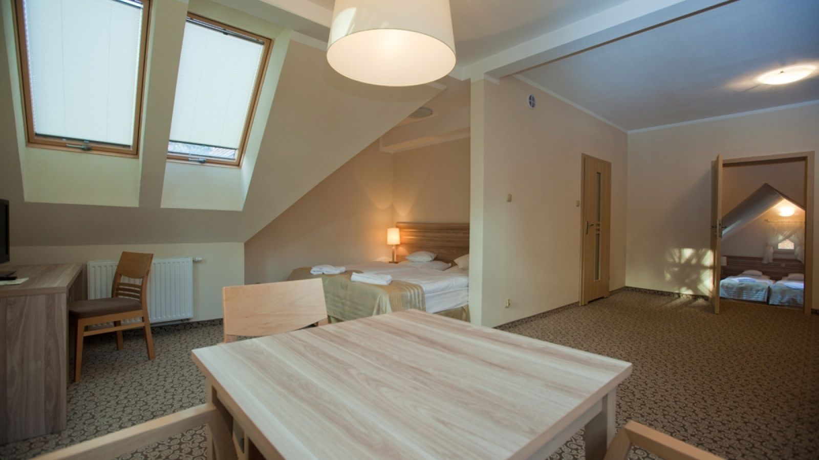 A room with a desk, two chairs, a bed, and a table. There are windows and a door.