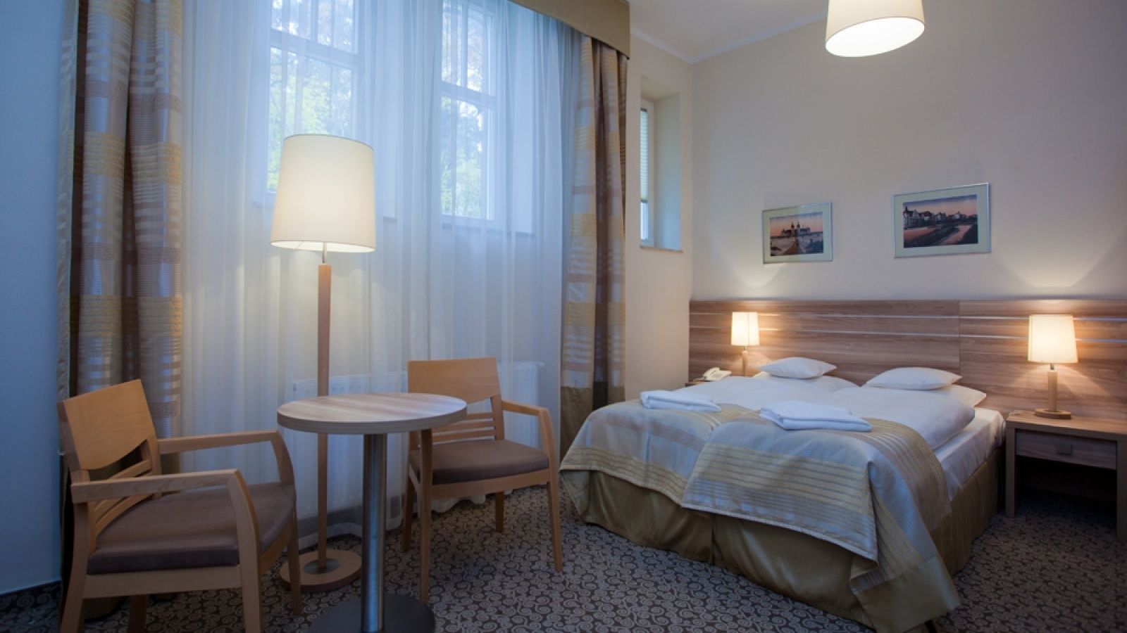 Habitación de hotel con cama, dos mesitas de noche con lámparas, una mesita redonda con dos sillas y una ventana con cortinas traslúcidas.