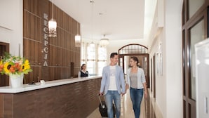 A reception area with a wooden reception desk, a receptionist, and a couple walking down a hallway with a modern interior design. - Hotel Cesarskie Ogrody (Swinoujscie)