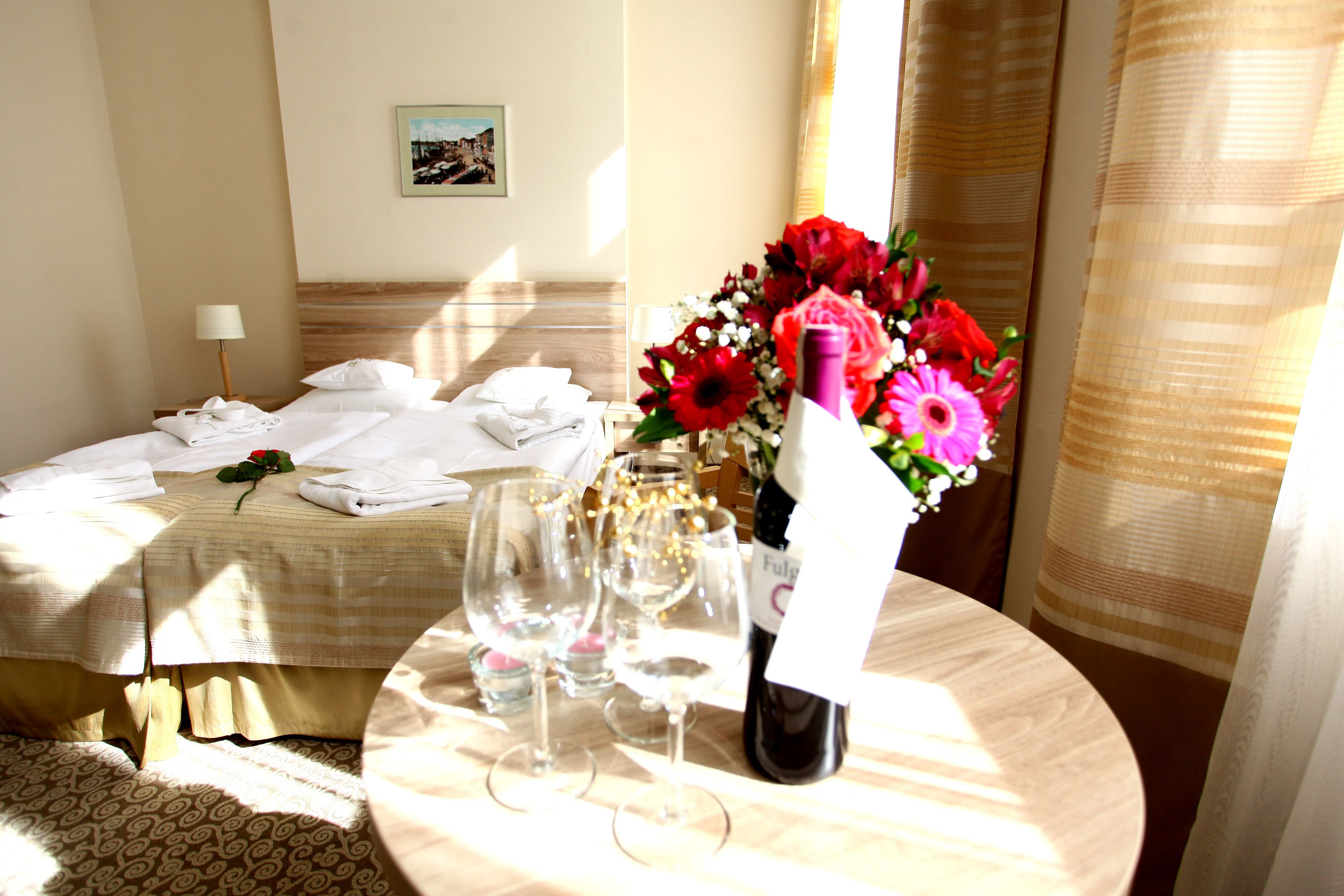 A hotel room with a neatly made bed, a table with wine glasses, a bottle of wine, and a bouquet of flowers.