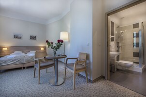 A hotel room with a bed, two chairs, a table with a vase of flowers, a lamp, and a shower area.