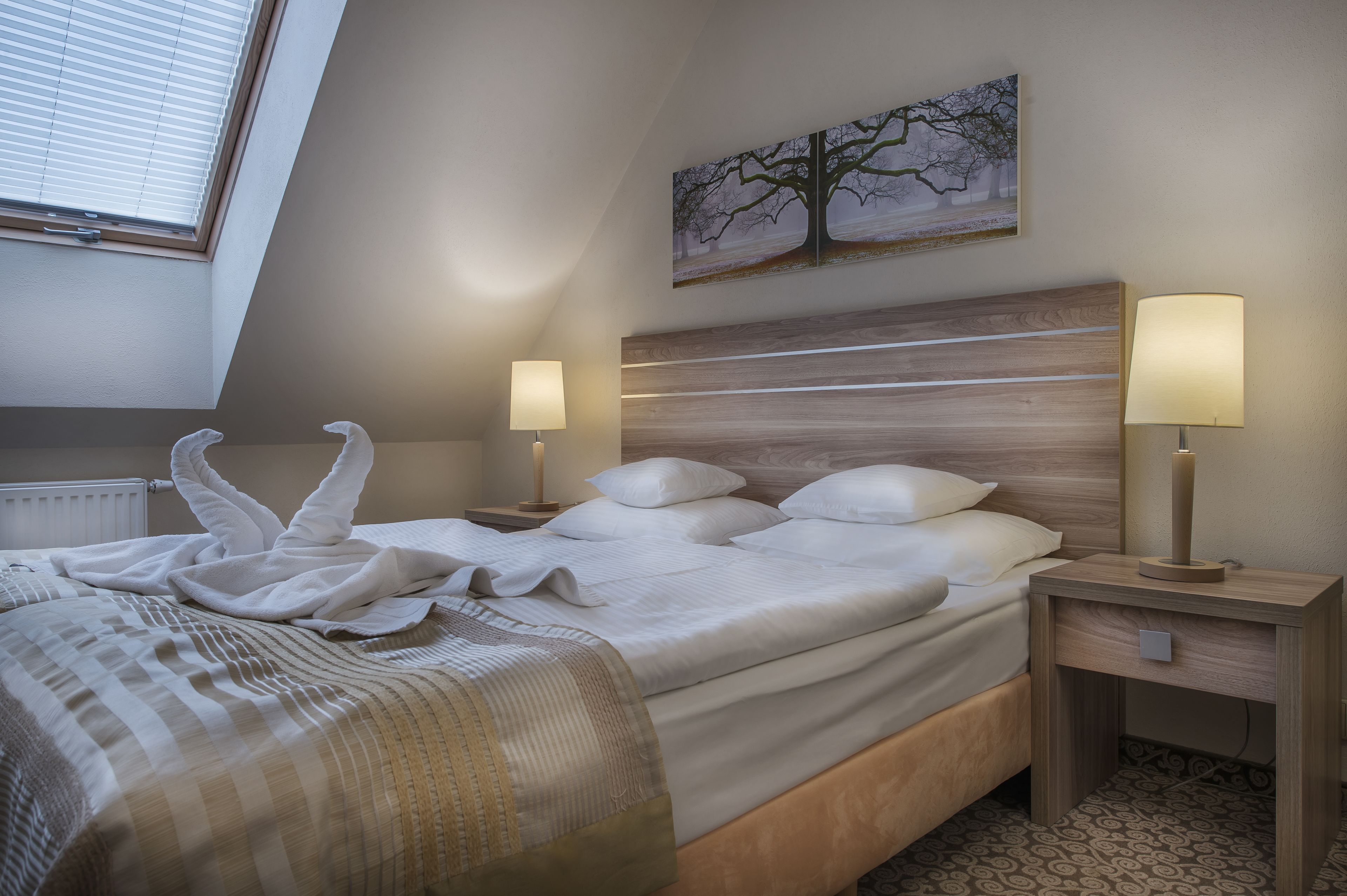 Una habitación de hotel con una cama, dos mesitas de noche con lámparas, un cuadro en la pared y una ventana con persianas.