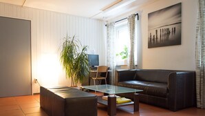 A living room with a glass coffee table, a leather sofa, a black ottoman, a potted plant, a TV, and a painting on the wall. - 1/2/sleep Hostel Nürnberg Messe (Nuremberg)
