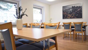 A dining area with a wooden table and chairs, a vase with foliage, and a cityscape painting on the wall. - 1/2/sleep Hostel Nürnberg Messe (Nuremberg)