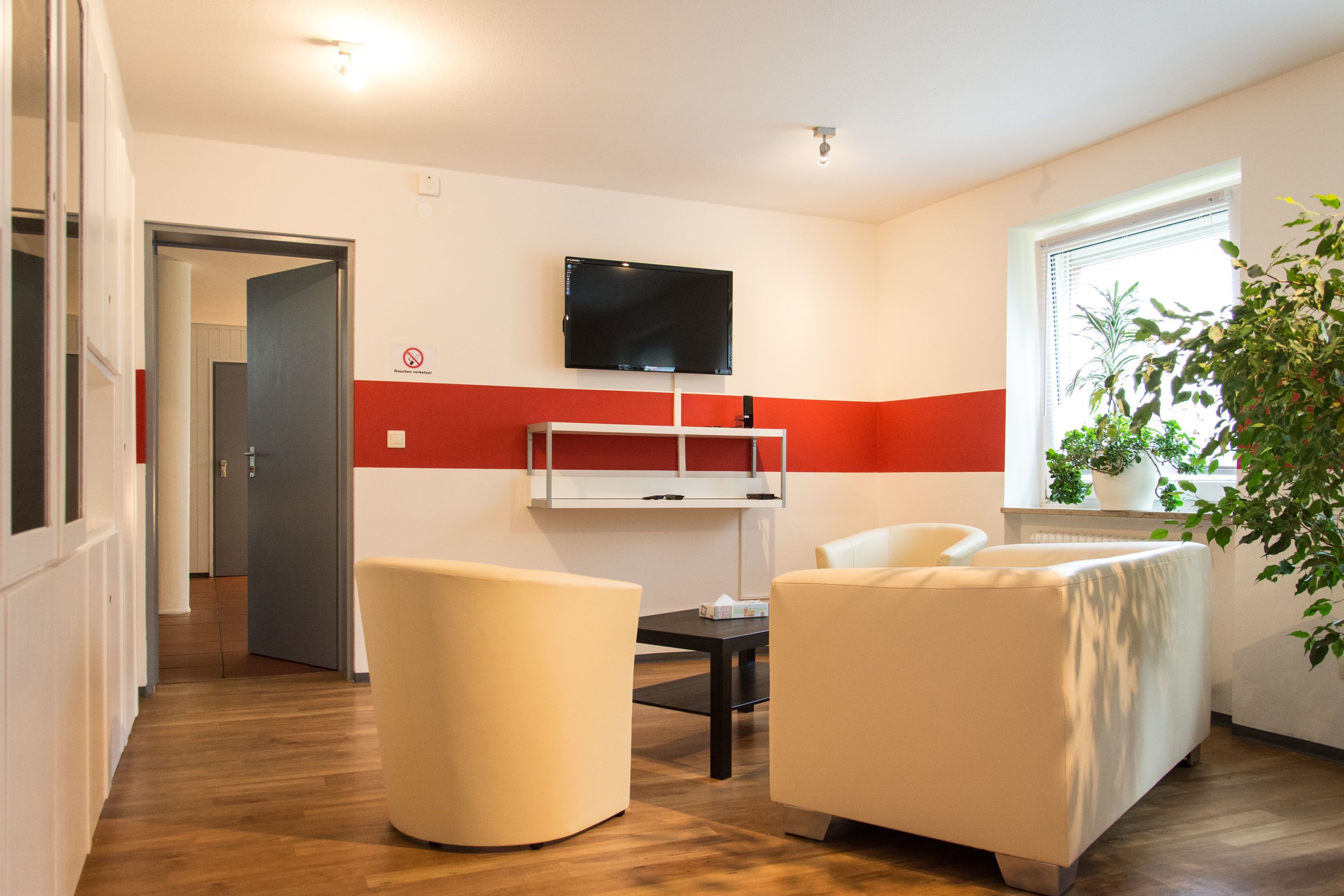 A modern hotel room with a wooden floor, a flat-screen TV mounted on the wall, a small table, two white armchairs, and a potted plant by the window.