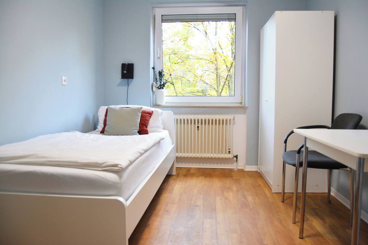 A bedroom with a bed, a radiator, a window with a view of trees, and a desk with a chair.