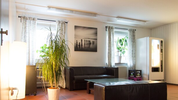 A living room with a sofa, a coffee table, a potted plant, a radiator, and a refrigerator. - 1/2/sleep Hostel Nürnberg Messe (Nuremberg)