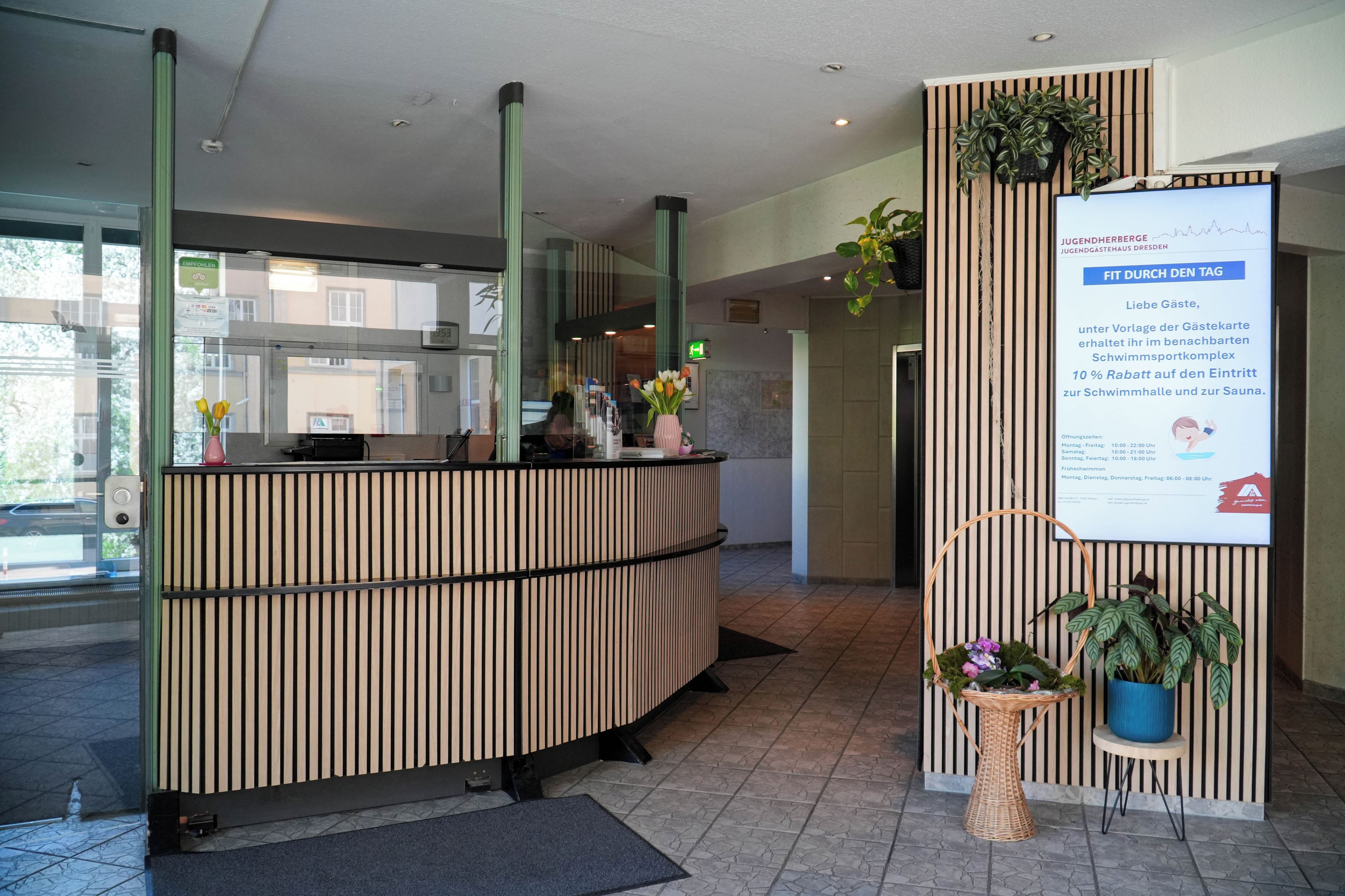 Un mostrador de recepción con diseño moderno, un panel informativo digital y plantas en macetas.